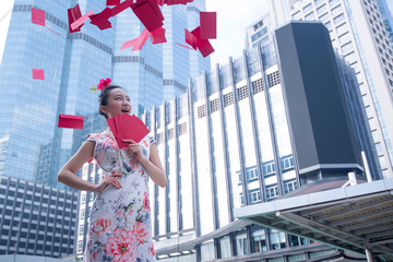 Excited Asian woman in Chinese dress smiling against modern building in city, Ang Pao red envelopes in hand and flying in the air, Chinese new year concept