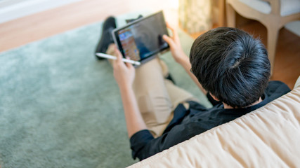 Fototapeta na wymiar Young Asian business man using digital tablet and pen while sitting on carpet by the bed in cozy bedroom. Home living lifestyle with modern electronic gadget concept