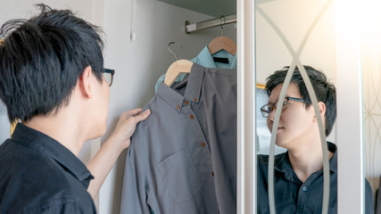 Young Asian man choosing casual style shirt in closet for dressing up in the bedroom. Home living lifestyle concept