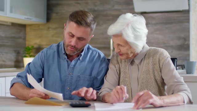 Medium shot of adult man and his aged mother sitting together at kitchen table and making calculations of family income