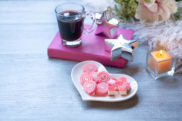 Sweet jelly in heart shaped plate for valentine's day, with candle, silver star, pink book and white fur on wood background