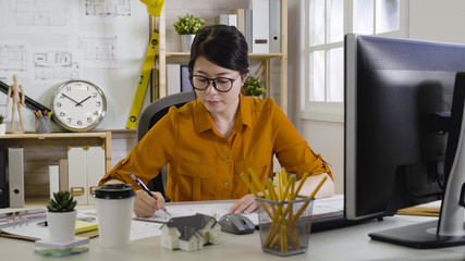professional young asian female architect wearing casual shirt sitting in cozy office and making...