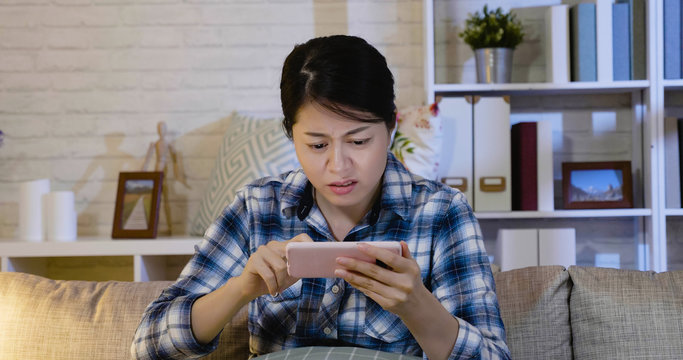 Worried Asian Woman Making Mistakes On Smart Phone Sitting On Sofa At Home At Night. Young Girl With Frowning Face Touching Cellphone Screen To Solve Problem. Confused Lady In Couch Angry With Device