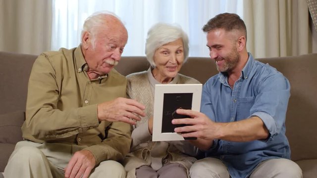 Medium shot of adult man sitting with his senior parents on sofa, giving them photo frame as present, then they rejoicing together
