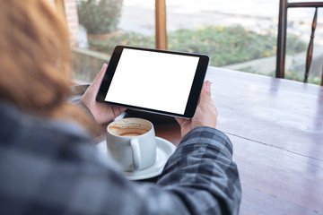 Mockup image of a woman holding black tablet pc with blank screen horizontally with coffee cup on wooden table in cafe