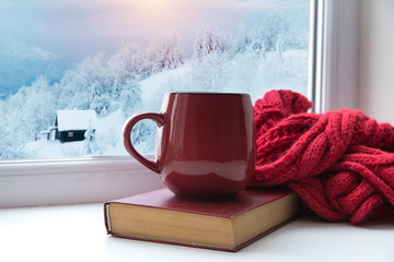 Winter background - cup with candy cane, woolen scarf and gloves on windowsill and winter scene outdoors. Still life with concept of spending winter time at cozy home with cold weather outdoors