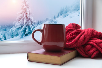 Winter background - cup with candy cane, woolen scarf and gloves on windowsill and winter scene outdoors. Still life with concept of spending winter time at cozy home with cold weather outdoors