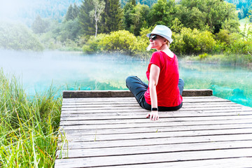 Girl with sunglasses and white hat sitting and enjoying on a wooden pier by the turquoise blue lake covered with light fog, Zelenci, Slovenia