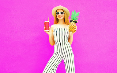 Happy smiling woman with pineapple, cup of juice in summer round straw hat, white striped jumpsuit on colorful pink wall background