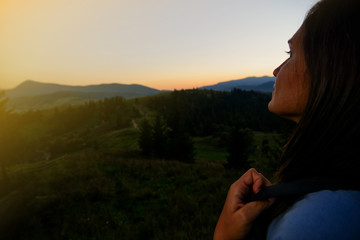 A young girl (tourist, traveler) with a backpack rejoices on top of the hills at sunset.