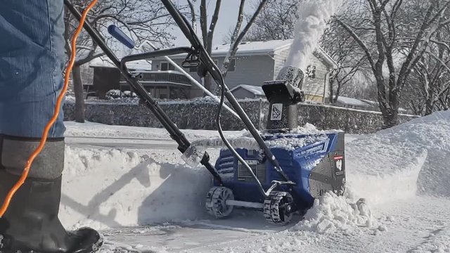 Close up and super slow motion of electric corded snowblower throwing snow while man ion jeans and boots pushes machine through pile of snow on his driveway.