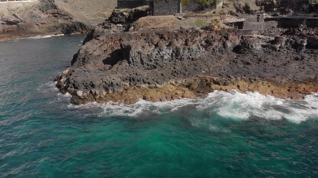 Aerial shot. Approaching the volcanic coast on which two unrecognized tourists take pictures. The landscape is big stones of yellow and brown color and beating about them turquoise waves on the beach