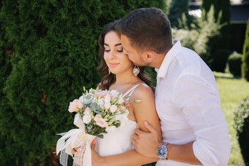 Handsome bearded man kiss his wife. Gogeous woman in white dress with beautiful bouquet. Stylish man in white shirt