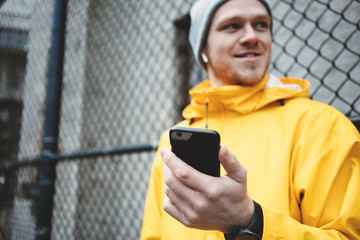 Smiling man in yellow raincoat standing on the street with mobile phone and wireless earphones