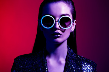 Fashion girl with long hair and round sunglasses in a black shining dress poses in neon light in the studio