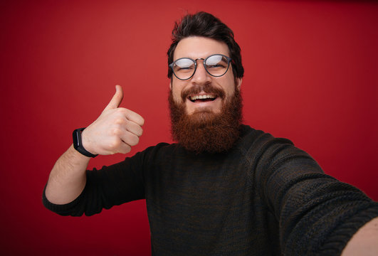 Happy bearded  man withsmart face making selfie and showing thumb up over red background