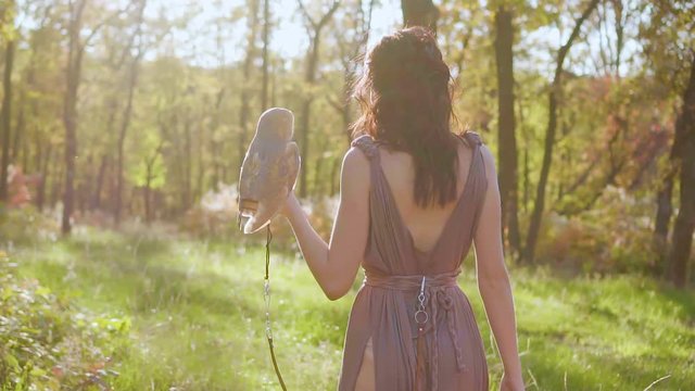 no face, greek goddess with dark curly hair walks in green garden, forest nymph in long brown pussy pale dress, lady with white barn owl on her arm, fairy of spring and animals in bright sunlight