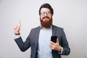 Bearded man wearing glasses and jacket holding mobile phone while pointing up with finger over gray background