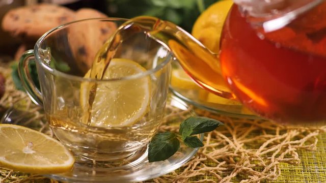 Tea being poured into glass tea cup with slice of lemon and mint leaf on saucer
