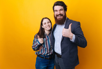Two smiling happy businesspeople in formalwear showing thumbs-up on yellow background