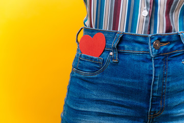 Close up on female wearing jeans with red heart in her pocket over yellow background - Powered by Adobe
