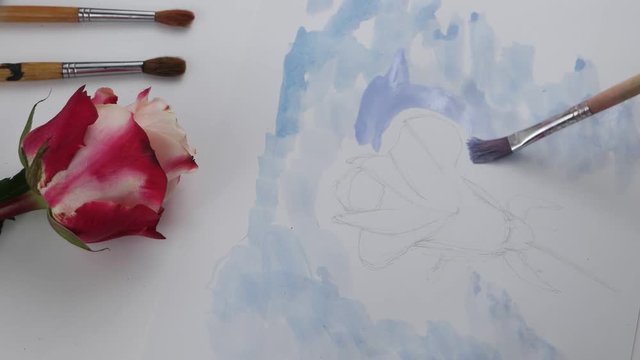 Drawing of background by blue gouache on the white sheet of paper on the background of paintbrushes and pink rose