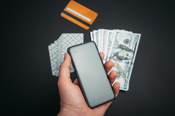 handsome hman hand holding smartphone with blank screen, playing cards and money on black background.