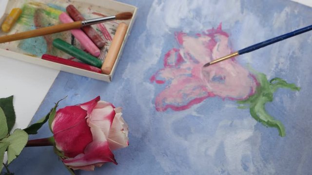 Drawing pink flower by pale pink gouache on the blue plywood on the background of pastels and pink rose