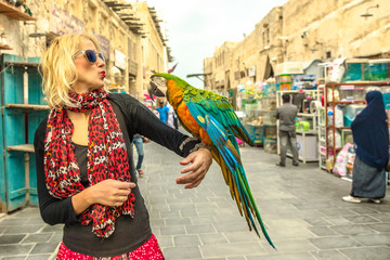 Happy woman kisses a parrot at Bird Souq inside Souq Waqif, the old market and tourist attraction in Al Souq District, Doha city center, Qatar. Caucasian traveler visits Middle East, Arabian Peninsula