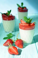 Italian dessert Panakota with strawberry coolies, fresh berries and mint on a blue background with hard shadows. Creamy milk tender dessert in a glass. Close up. Vertical shot. Summer dessert. 