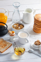 Healthy breakfast eating concept, various morning food - pancakes, soft-boiled egg, toast, oatmeal, granola, fruit, coffee, tea, orange juice, milk on white wooden table