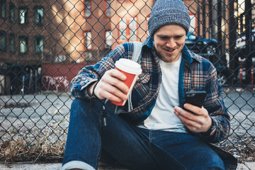 Handsome man sitting on the city street resting with cup of hot coffee and using mobile phone
