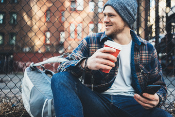Handsome man sitting on the city urban street resting with cup of hot coffee and using mobile phone