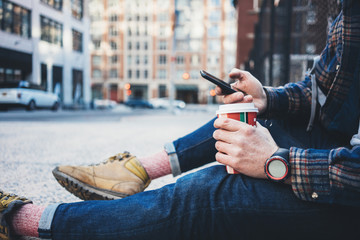 Casual man sitting on the street sidewalk resting with cup of hot coffee and using mobile phone. Close-up