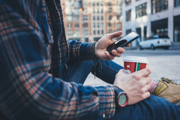 Man sitting on the street sidewalk resting with cup of hot coffee and using mobile phone. Close-up