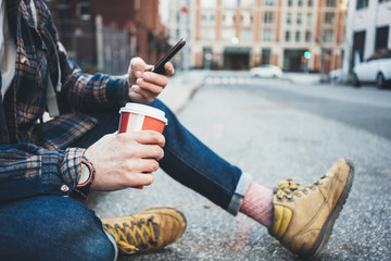 Casual man sitting on the street resting with cup of hot coffee and using mobile phone. Close-up