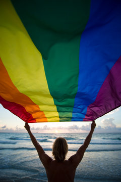 Scenic silhouette of a man with blond hair holding a gay pride rainbow flag blowing in the wind on a tropical beach with golden sun