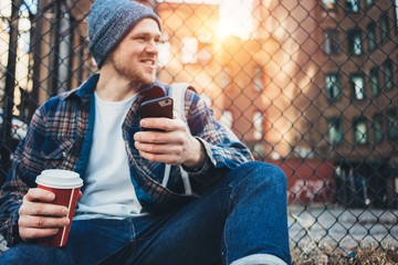 Smiling man sitting on the city street resting with cup of hot coffee and using mobile phone