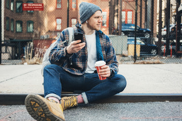 Handsome man sitting on the city street resting with cup of hot coffee and talking on mobile phone using wireless earphones