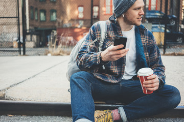 Casual man sitting on the city street resting with cup of hot coffee and talking on mobile phone using wireless earphones