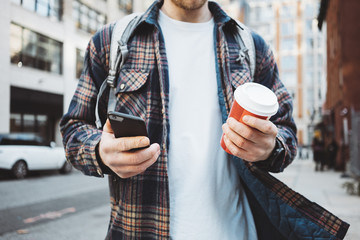 Casual man walking on the urban street sidewalk with cup of hot coffee and texting on mobile phone. Close-up