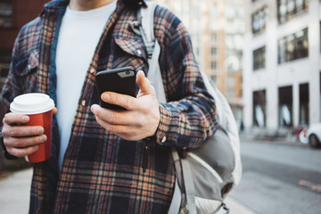 Casual man walking on the street sidewalk with cup of hot coffee and texting on mobile phone. Close-up