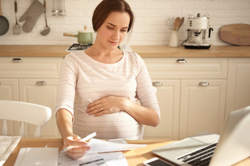 Indoor shot of busy serious young pregnant woman keeping one hand on belly, holding sheet of paper,...