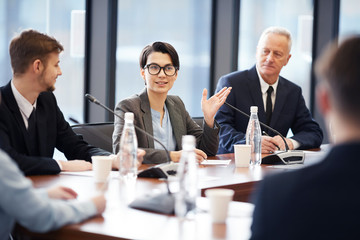 Portrait of young businesswoman speaking to microphone during group discussion in conference room,...