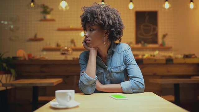 beautiful sad african american female customer propping chin with hand while waiting for someone in cafe