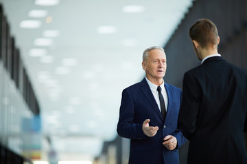 Waist up portrait of successful senior businessman talking to young employee standing in hall of...