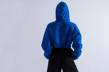 Back photo of an anonymous  girl in blue hoodie with hood on and pants. Hands in pockets. Sport casual style with copy space. Closeup on white background.