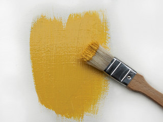A small paint brush with yellow paint lies on a white surface with fresh yellow paint strokes.