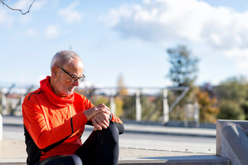 Senior runner man sitting and looking at smartwatch during training. He is satisfied with results.