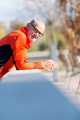 Front view of one senior runner man with sportswear sitting on a wooden fence while looking camera